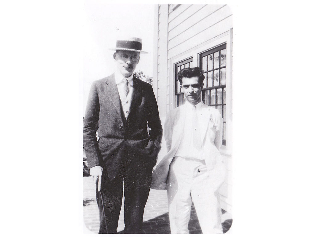 Two Fellows stand next to one another outside. They are both dressed in suits.