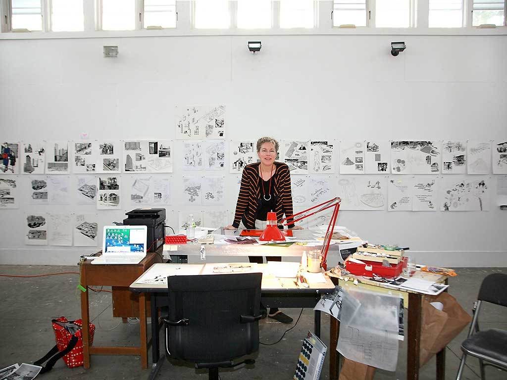 Fellow Julia Lacquette stands at a desk in her studio. On the wall behind her are many sketches and drawings