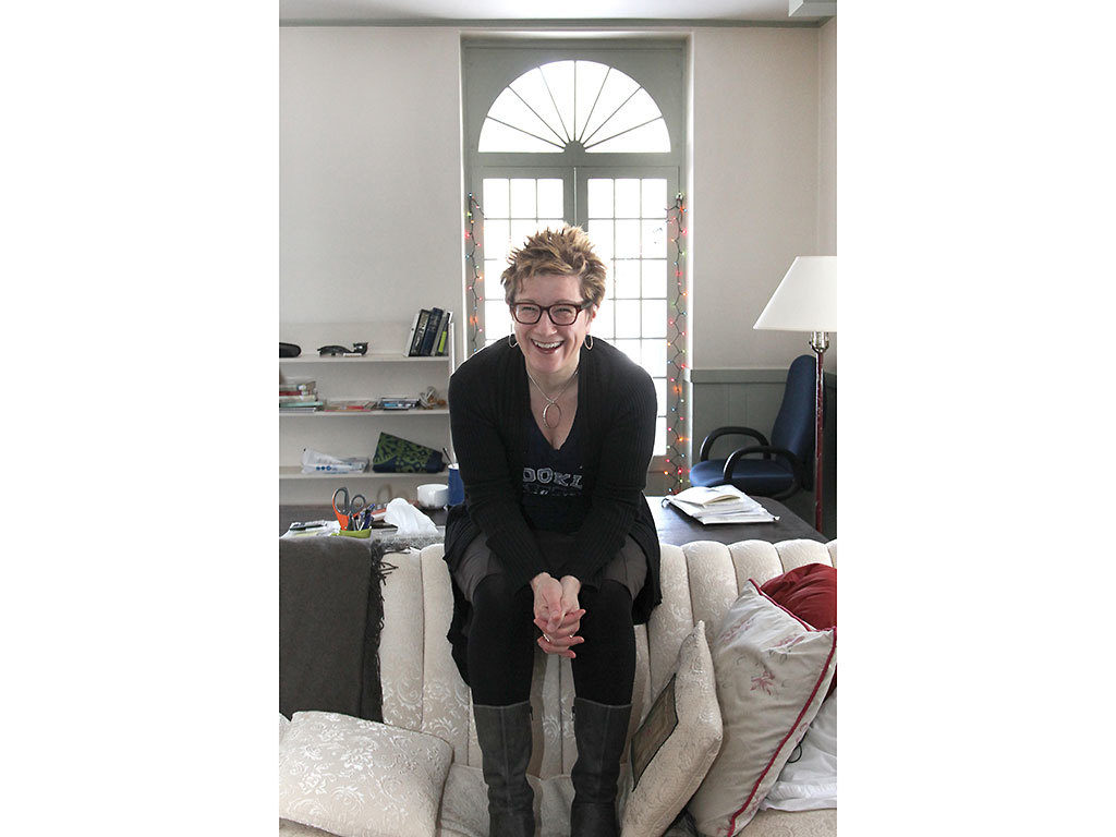 Portrait of Fellow Lisa Kron in her studio. She is sitting on the back of a couch and smiling