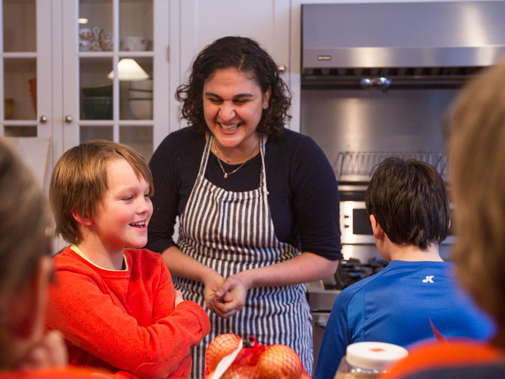 A woman in a kitchen with a group of children. They are all cooking together and laughing