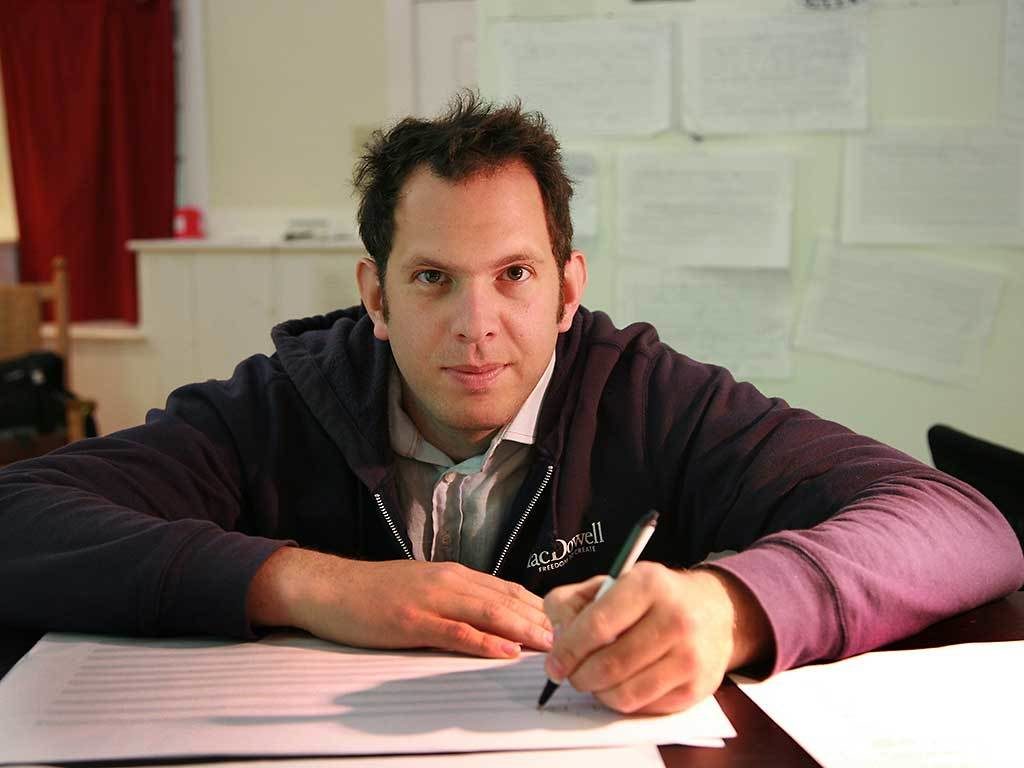 Fellow Yotam Haber writing on a piece of paper and looking straight at the camera.