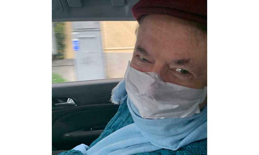 A man wearing a face mask smiles at the photographer while sitting in a car
