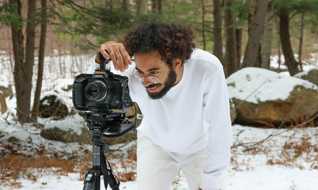 Filmmaker Juan Luis Matos is dressed in all white in MacDowell's woods with an early season snowfall covering the ground. He is looking at the composition screen of his tripod-mounted camera.