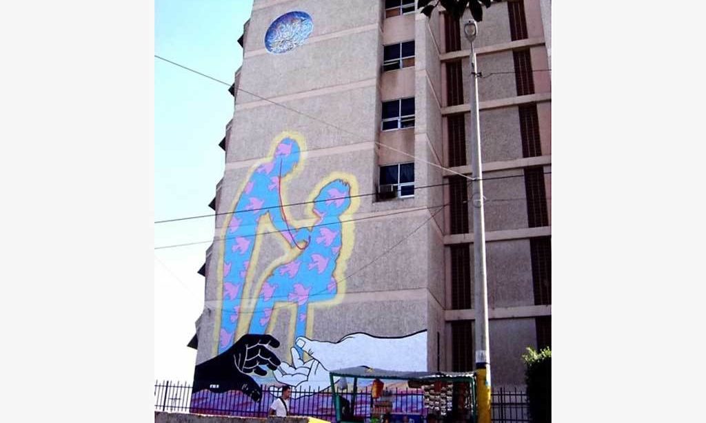 A mural on a tall building taken from far away. Two large silhouetted figures are at the top. Below them are two hands reaching toward one another. Underneath it all is a scene with a purple sky and many, small, simply drawn figures.