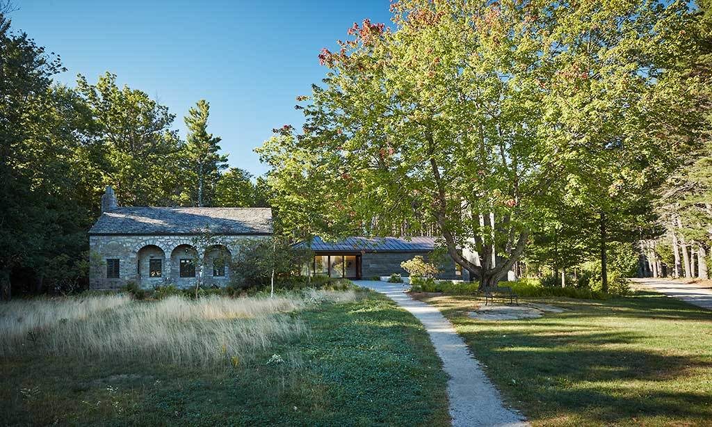 MacDowell's Library from a distance. A dirt path leads to the building. A lush meadow on the left, large deciduous trees on the right.