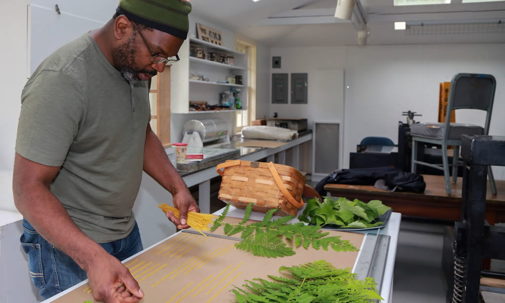 Fellow Paul Rucker stands at a desk. He is arranging fern leaves for a project