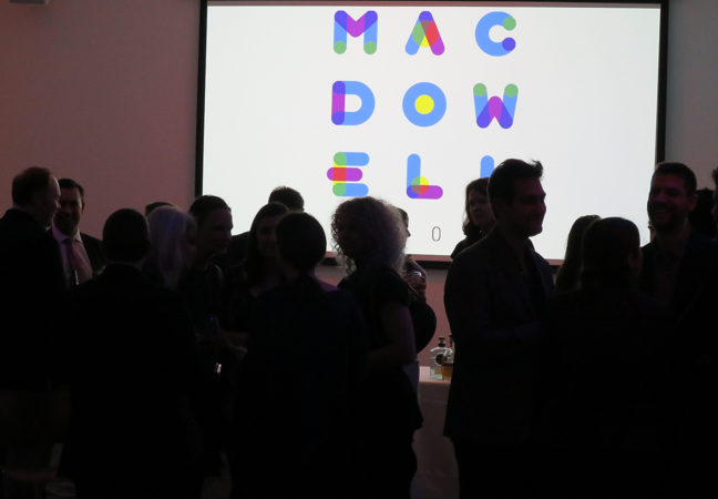 A group of people are silhouetted against a large screen in a dark room. The screen is showing the MacDowell logo