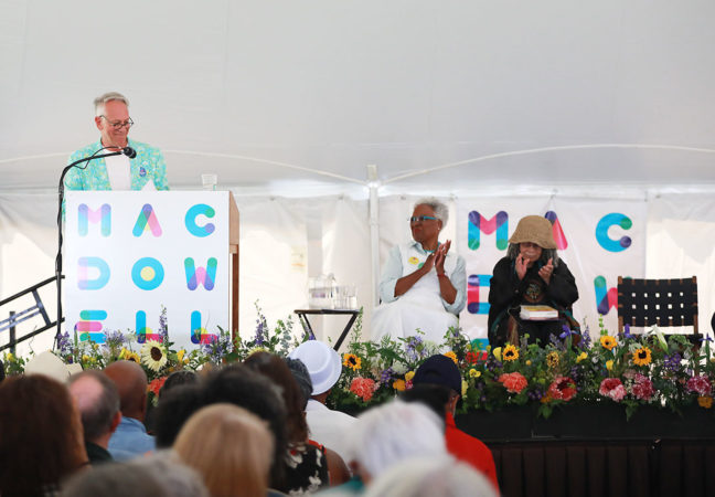 MacDowell Executive Director Philip Himberg speaks from the Medal ﻿Day stage on July 10, 2022. Crowd can be seen in foreground, as well as other VIPs, Chair Nell Painter, Medalist Sonia Sanchez, and introductory speaker and Fellow Walter Mosley are seated on the stage.