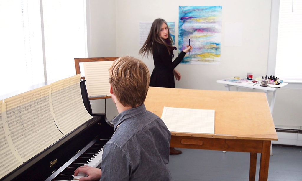 Two Fellows working together in a studio. The man sits at the piano and plays while the woman stands and paints with bright colors