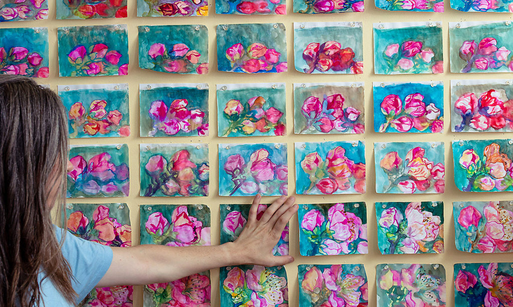 A Fellow stands in front of a wall covered in small paintings of flowers. The series of paintings are colorful, her hand rests against the wall gently.