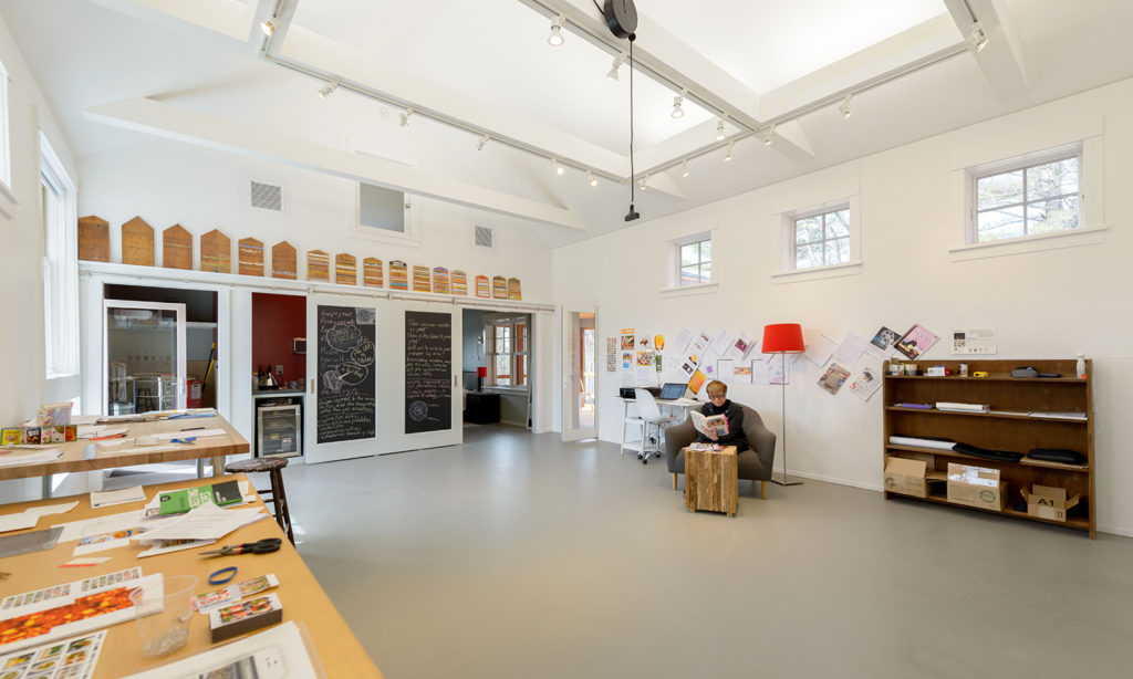 Interior of Eastman studio. The bright room is host to a Fellow who has their work and materials laid out on a table. The Fellow sits in an armchair on the far side of the room
