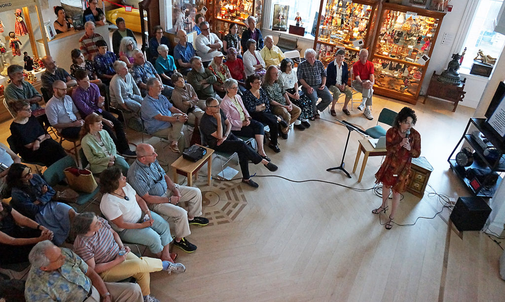 A Fellow stands at the front of the room as she presents to a small crowd. They are in the main space of the Mariposa Museum where there are glass display cases with artwork and artifacts lining the walls