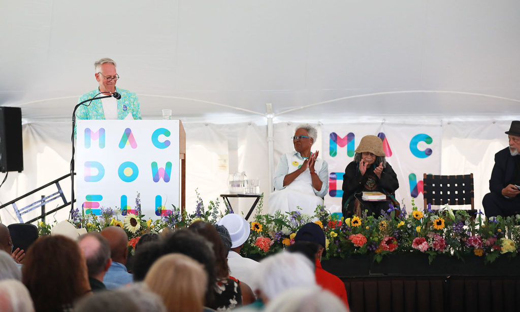 MacDowell Executive Director Philip Himberg speaks from the Medal ﻿Day stage on July 10, 2022. Crowd can be seen in foreground, as well as other VIPs, Chair Nell Painter, Medalist Sonia Sanchez, and introductory speaker and Fellow Walter Mosley are seated on the stage.