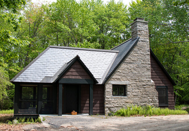 Delta Omicron studio in spring time. The dark-sided studio has a large stone chimney. The building is surrounded by a tall forest with bright green leaves, ferns begin to grow around it