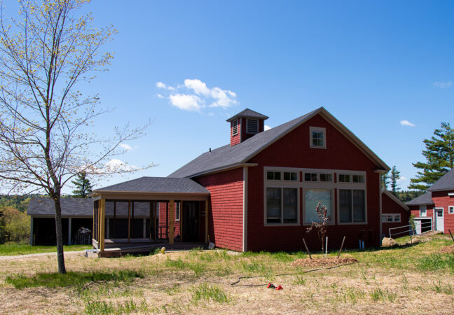 Firth studio in spring time. New grass and trees have been planted in front of the newly renovated studio, The large, red building sits on top of a hill and has a large screened in porch on its left side.