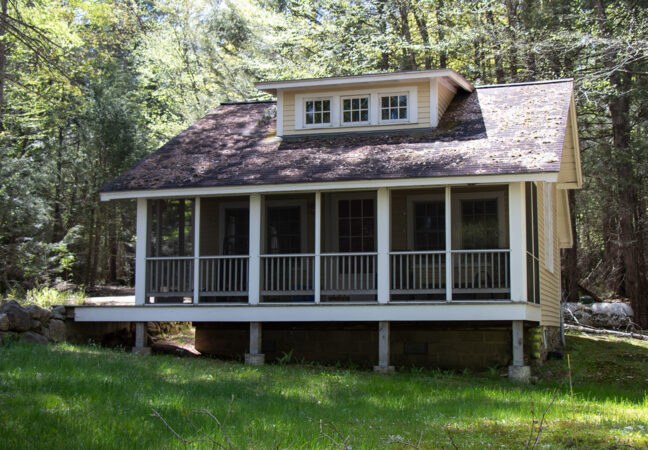 New Jersey Studio in spring time. In front of the large screened in porch is a lush, dark green lawn. The sun is shining brightly through the surrounding forest which is beginning to get its leaves