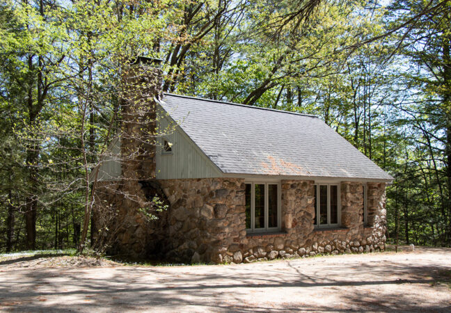 Sprague-Smith studio in the spring time. The small stone building is surrounded by trees starting to leaf-out and the ground is covered in the beginnings of lush ferns and grass.