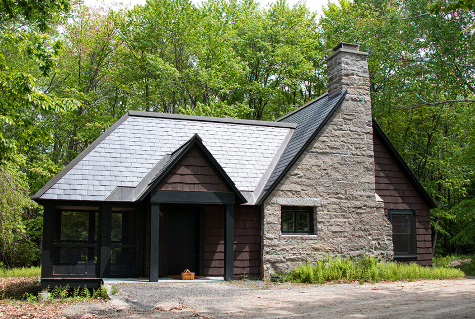 Delta Omicron studio in spring time. The dark-sided studio has a large stone chimney. The building is surrounded by a tall forest with bright green leaves, ferns begin to grow around it