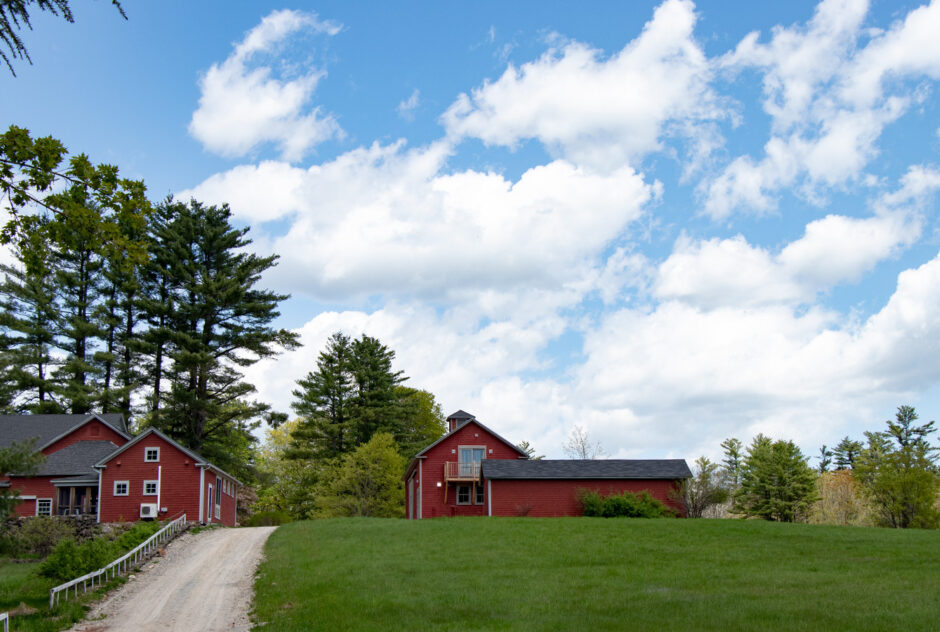 Firth studio in spring time. The red building sits at the top of a hill, a large field spread behind it. The field is a bright, lush green and there are fluffy white clouds in the bright blue sky. Another set of red buildings and studios are on the left side of Firth, across a dirt road.