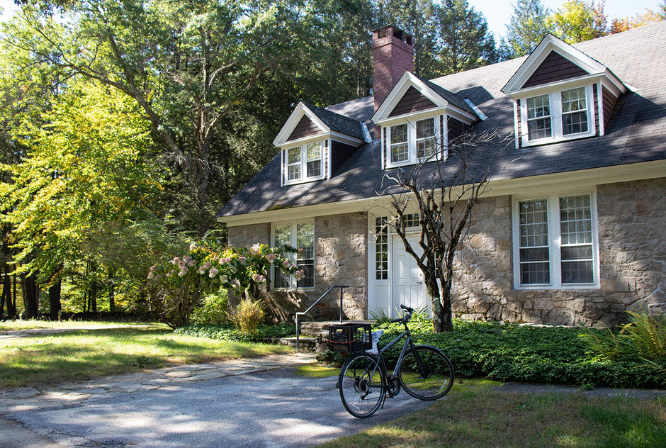 Banks studio in summertime. The stone building is surrounded by lush, green trees and a lawn. A bicycle sits outside the front door.