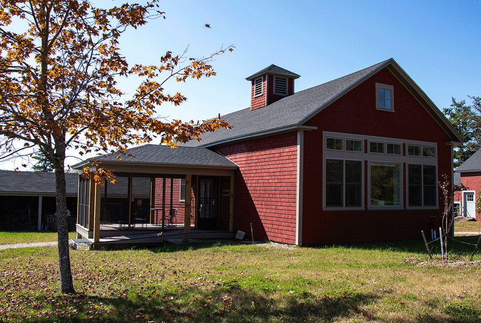 Firth studio in early fall. The red building sits atop a hill and a small tree is growing out front. A small screened porch sticks off the side of it. The sky is a clear blue and the sun is shining brightly.