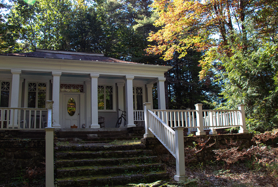 Watson studio in the fall. Seen from the studios courtyard, the building is nestled into a dense forest. Columns line the porch and a white fence circles the lawn. a sinking sun lights the scene.
