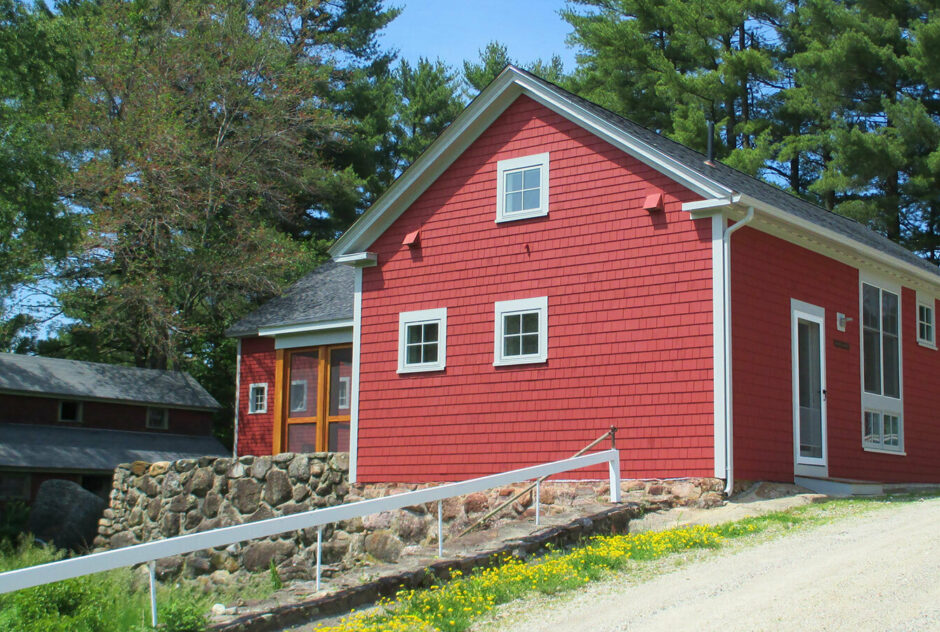 Eastman Studio in summertime. Seen from down the hill it sits on, the red building is supported by a stone foundation. Tall pine trees act as a backdrop to the sunny, blue-skied scene.