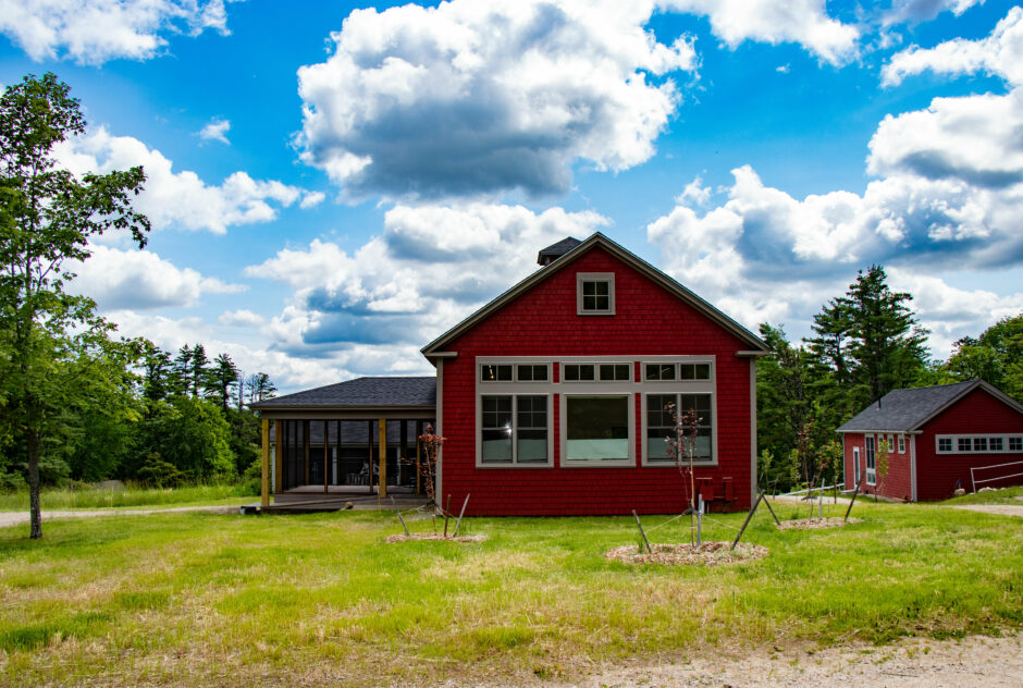 Firth studio on a bright sunny day in summer. There are fluffy white clouds littering the sky. The red building, with a screened porch off its side, sits atop a hill with a newly planted lawn out front.
