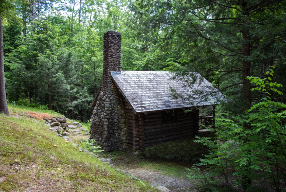 The log cabin in summer. The small log cabin, with its tall stone chimney, sits partway down a hill leading into a dense forest.