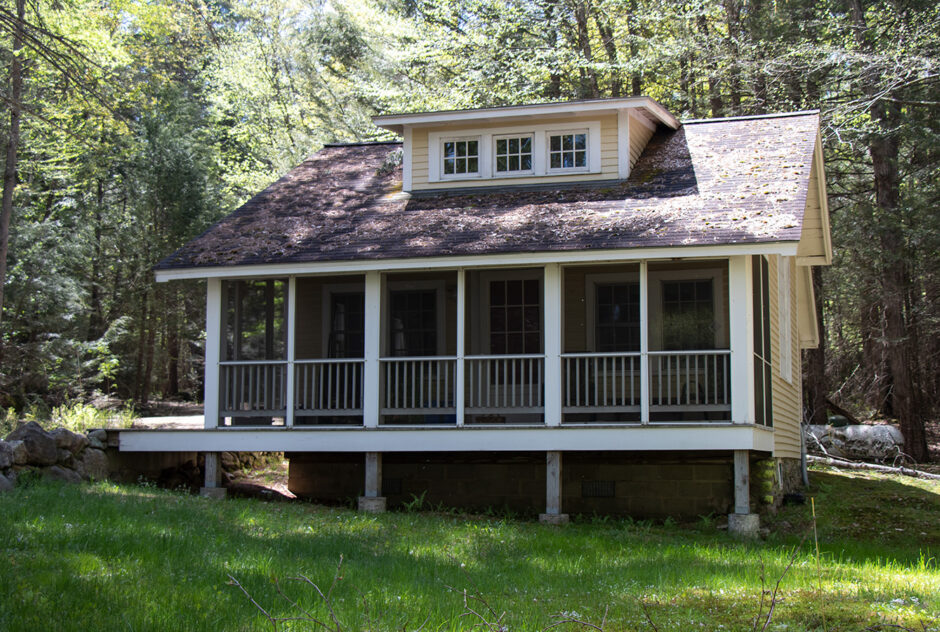 New Jersey Studio in spring time. In front of the large screened in porch is a lush, dark green lawn. The sun is shining brightly through the surrounding forest which is beginning to get its leaves